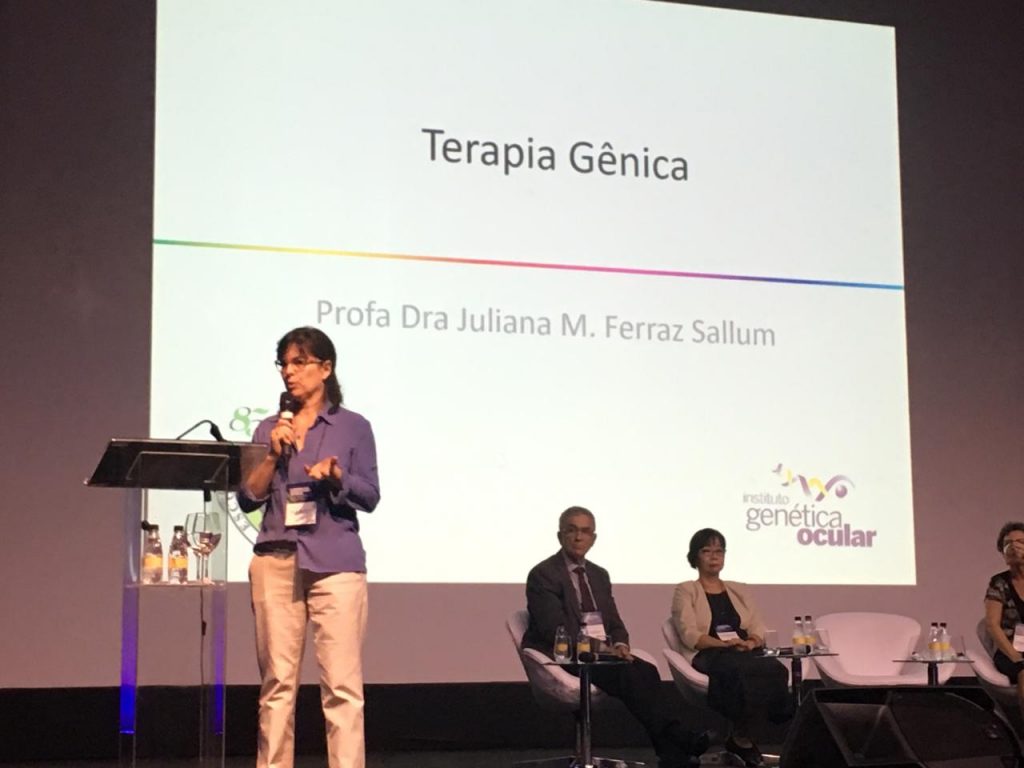 #PraCegoVer foto da Profa. Dra. Juliana Sallum palestrando no palco no evento. Ao fundo há uma projeção com o título "Terapia Gênica"
