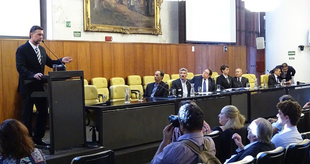 #PraCegoVer foto da mesa de abertura da Cerimônia. Na foto há um homem de pé discursando, e ao seu lado direito estão sentados em uma mesa comprida vários homens de terno. Todos eles estão de frente para a platéia, que não aparece na foto.