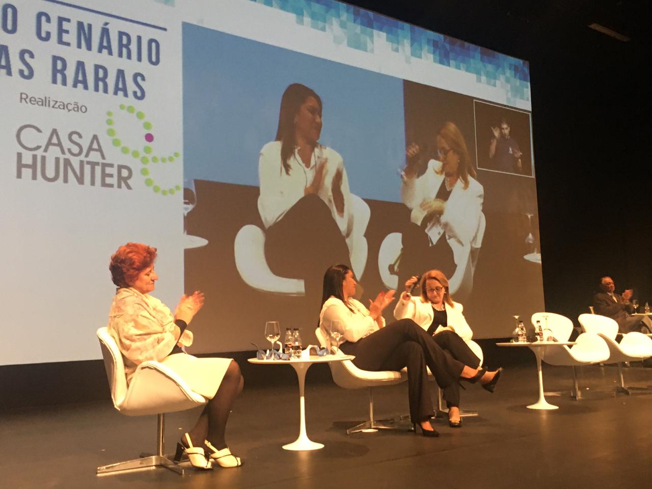#PraCegoVer fotografia do evento. A foto mostra o palco do evento, onde está ao lado de outras pessoas, a presidente da Retina Brasil, Sra. Maria Júlia Araújo. No momento da foto a Sra. Maria Júlia é aplaudida.