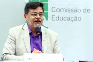 Foto de um homem branco, chamado Barbosa, de barba grisalha e cabelos pretos, de terno claro, camisa roxa, usando óculos. Atrás dele uma parede com o escrito: comissão de Educação